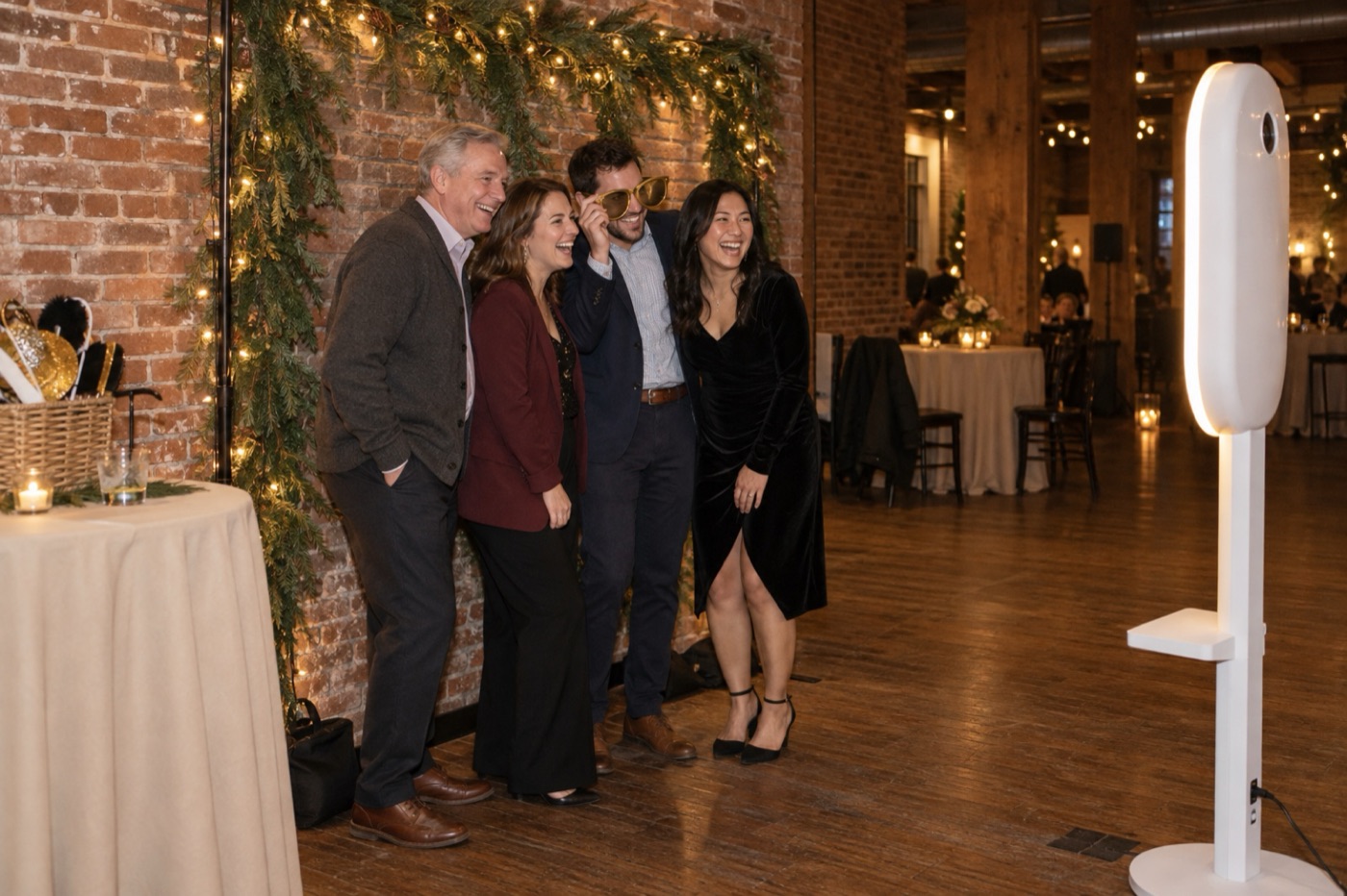 Four coworkers posing in front of an evergreen and string-light backdrop while the white photo booth faces them