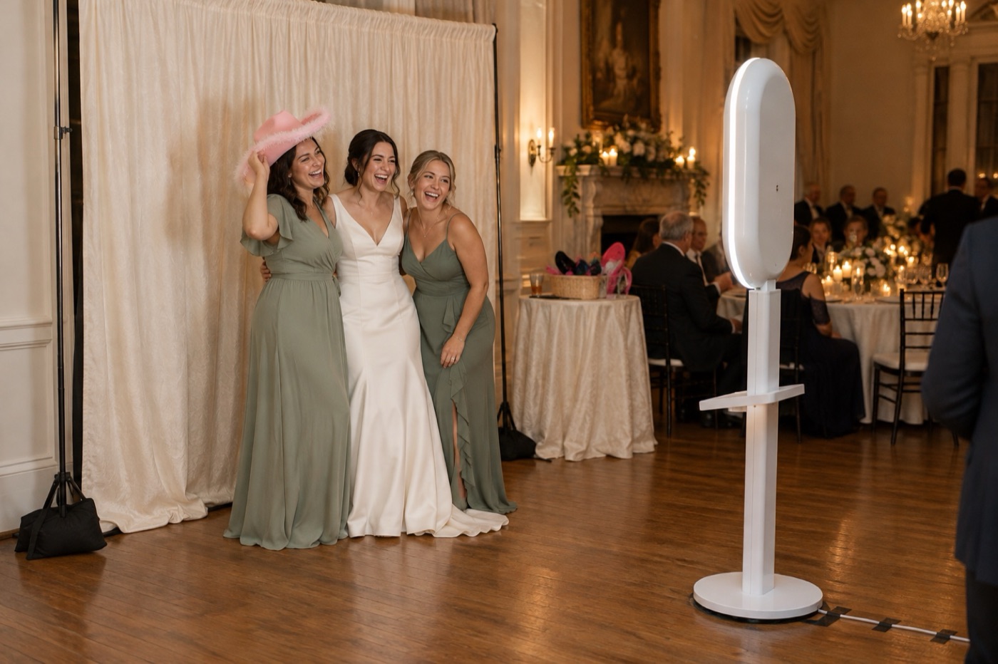 A bride and two bridesmaids posing in front of an ivory drape backdrop, facing the white photo booth